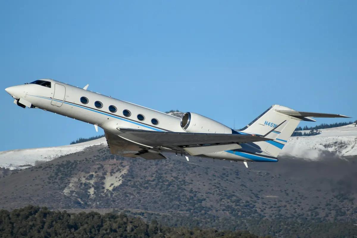 A business jet climbing past a snow-dusted mountain ridge under a clear blue sky
